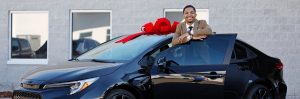 Young man posing with a new black car featuring a large red gift bow on the roof.
