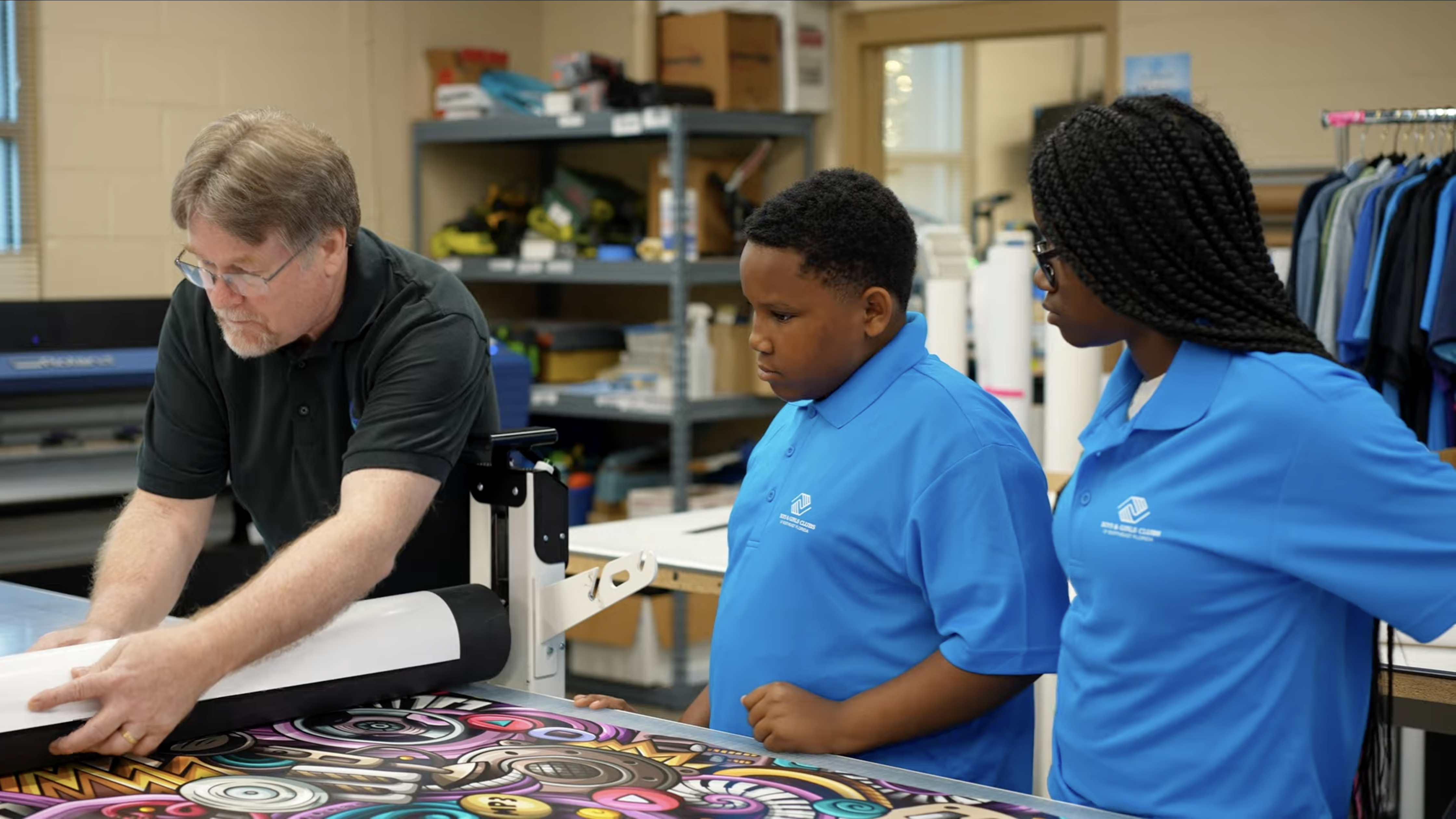 An instructor shows two students in blue polo shirts how to operate a vinyl printer.