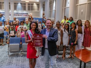 Miss Florida holding a plaque with a man in an auditorium surrounded by children and staff.