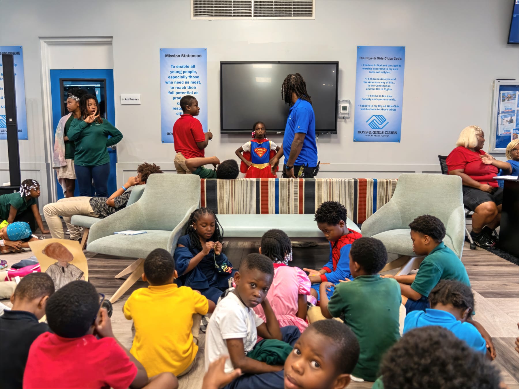 A group of kids sitting on the floor and surrounding furniture during an activity at a Boys & Girls Club facility with posters on the wall.