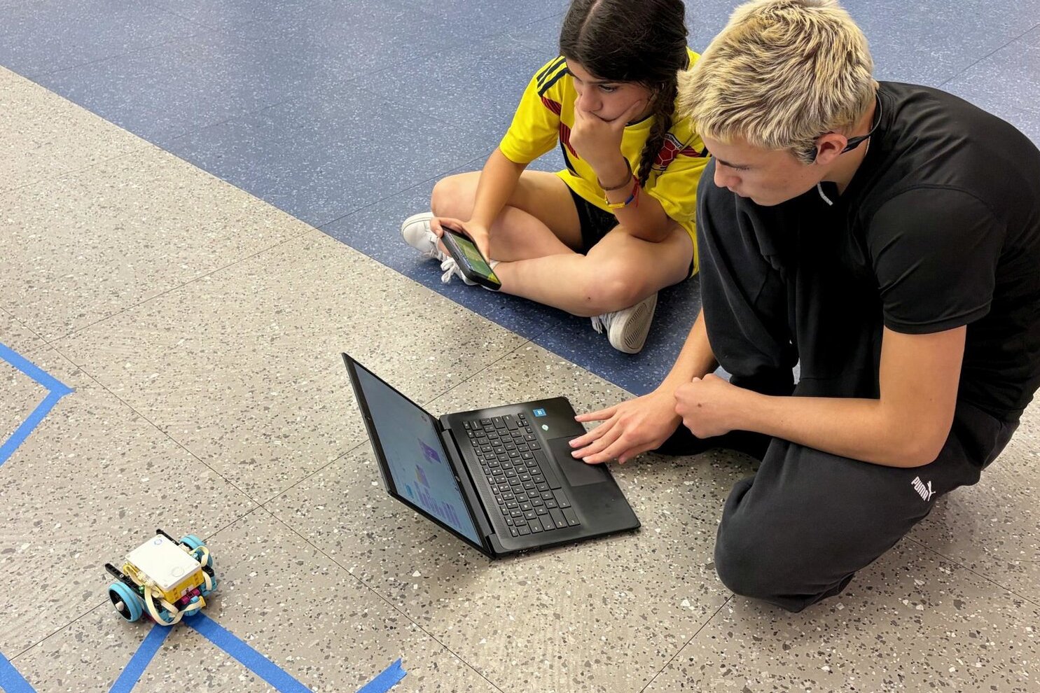 Two students sitting on the floor programming a small wheeled robot using a laptop.