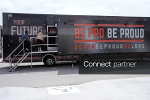 Two people entering a "Be Pro Be Proud" mobile workshop trailer parked outdoors.