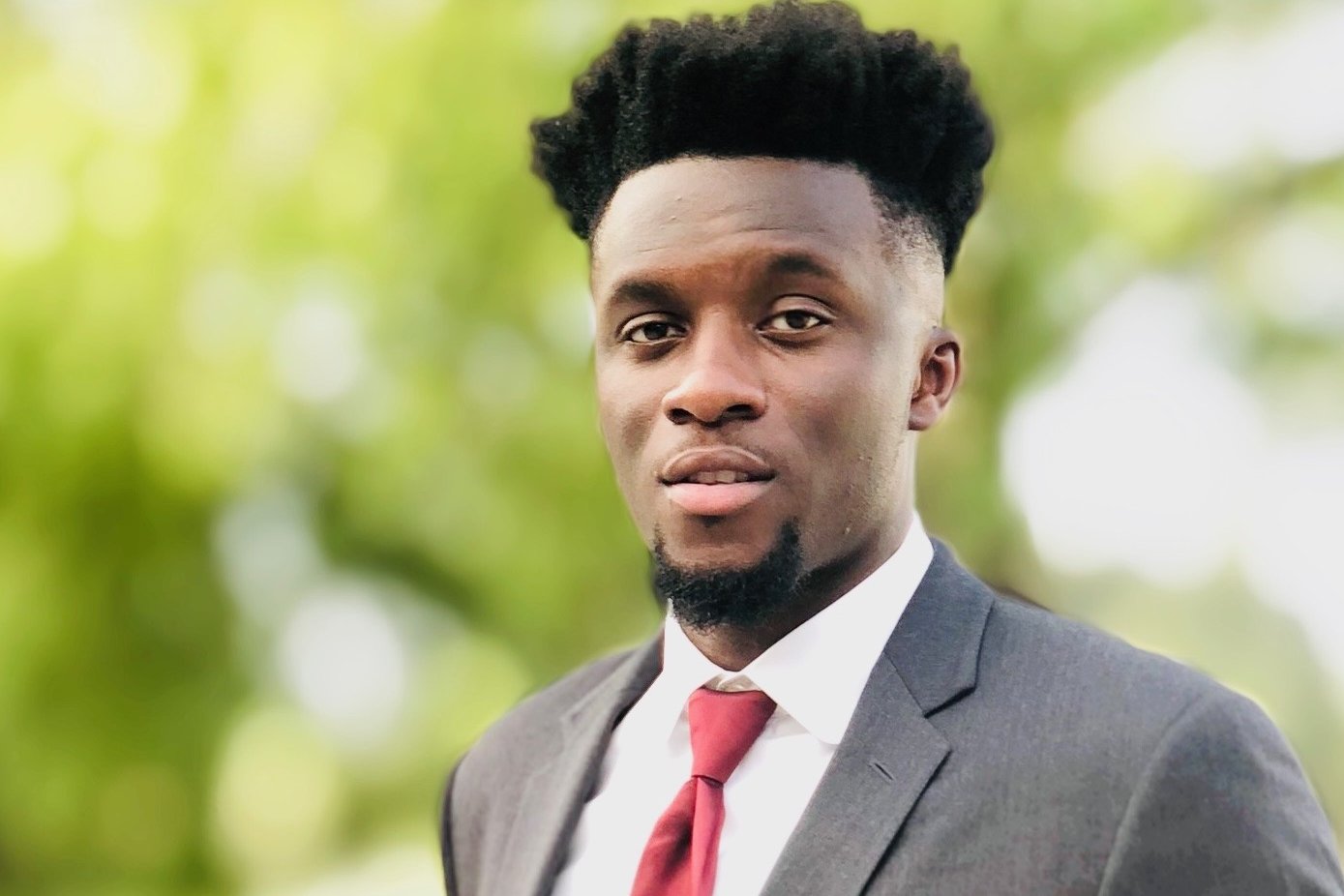 Professional headshot of a young Black man wearing a dark suit jacket, white dress shirt, and red tie outdoors.